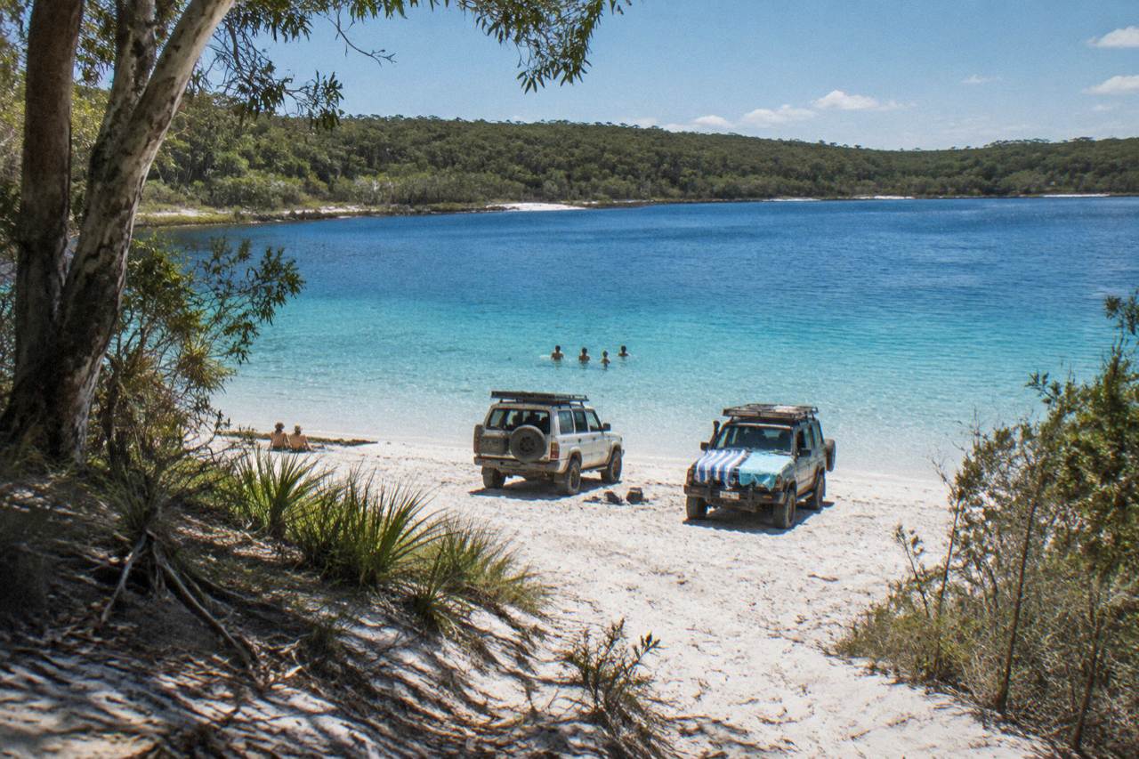 Four wheel drive tour vehicles parked on a wide sand beach beside turquoise lake water