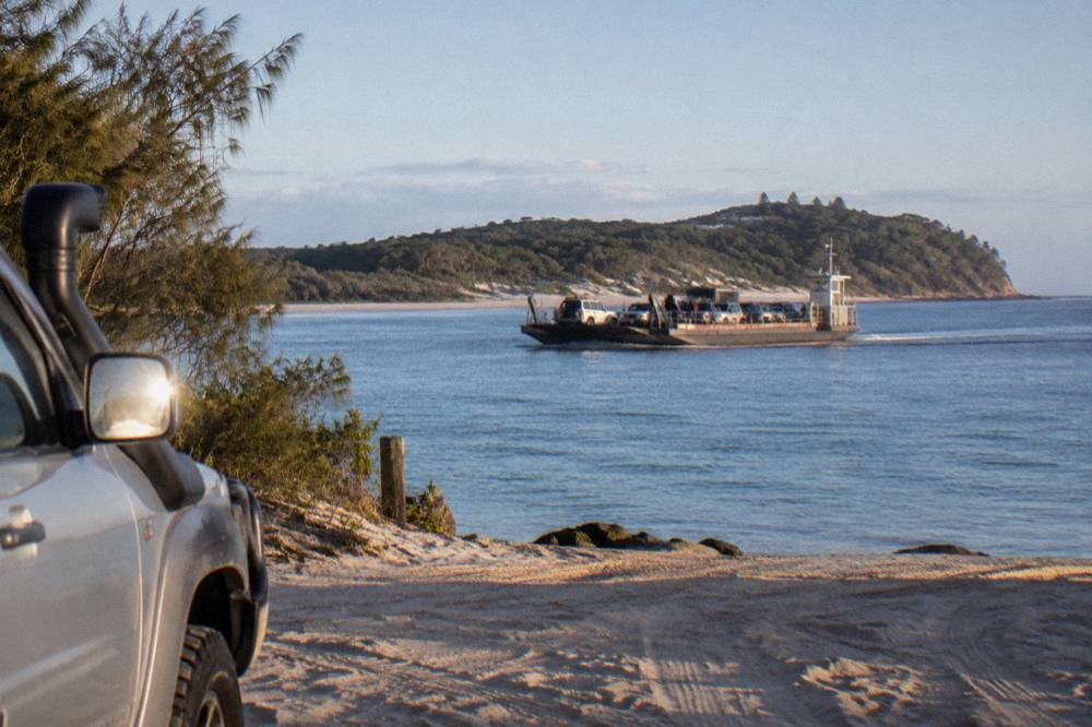 A vehicle barge crossing calm blue water toward a forested sand island
