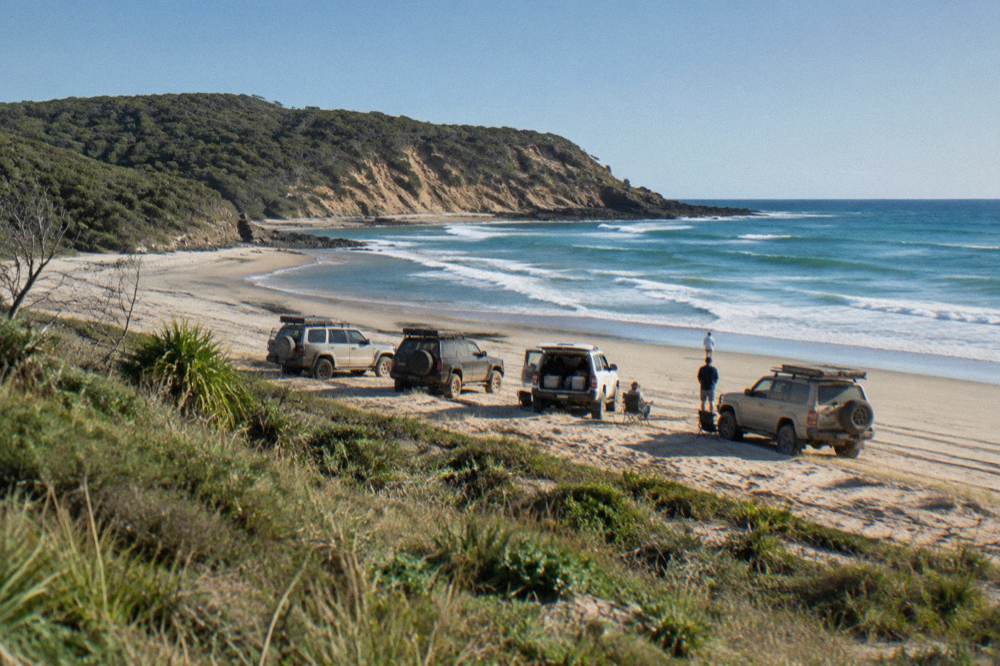 A convoy of four wheel drive vehicles parked together on a sandy beach beside a headland