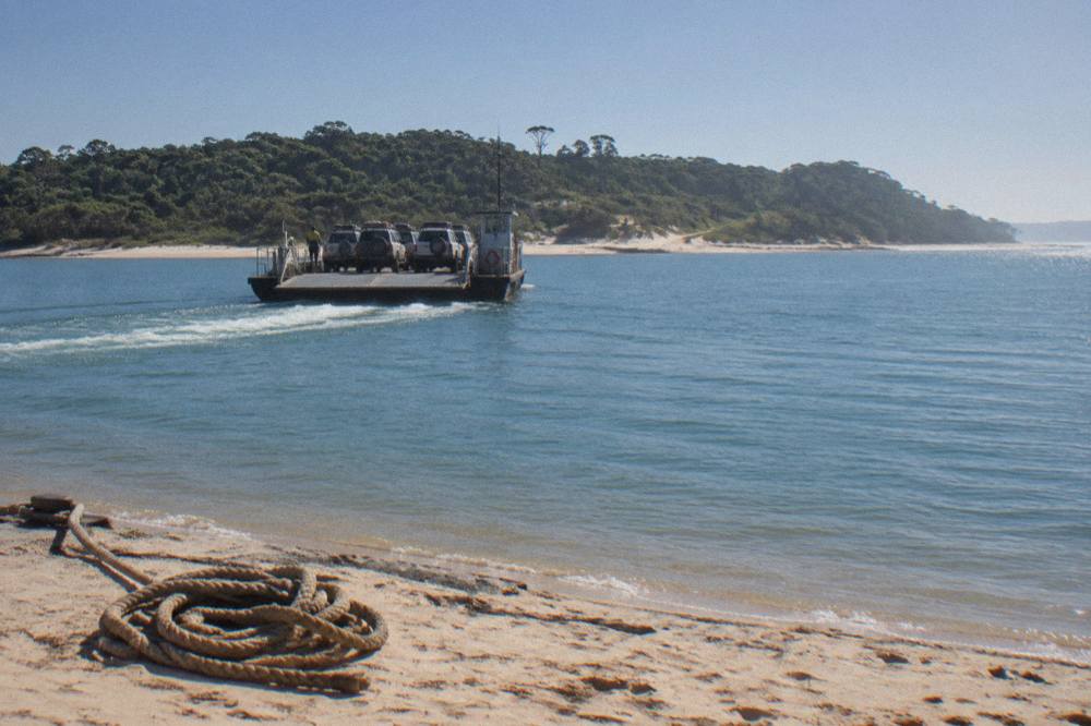 A vehicle ferry loaded with four wheel drives crossing calm blue water toward a tree covered sand island