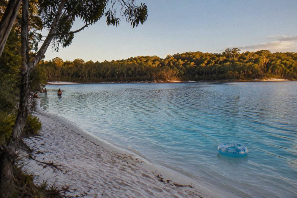 A clear freshwater lake ringed by white sand and tall eucalypt forest under a blue sky