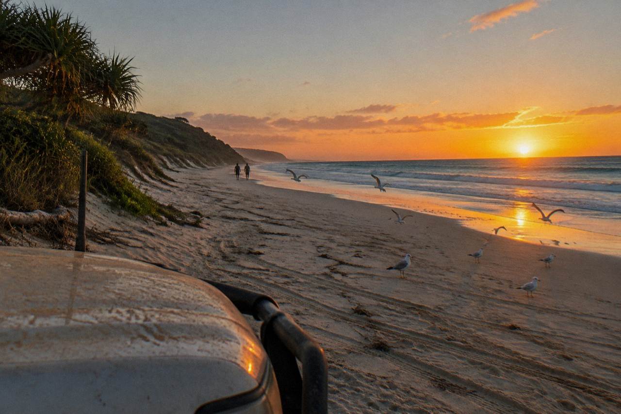 Four wheel drive vehicles parked on a wide sandy beach beside turquoise water at sunrise