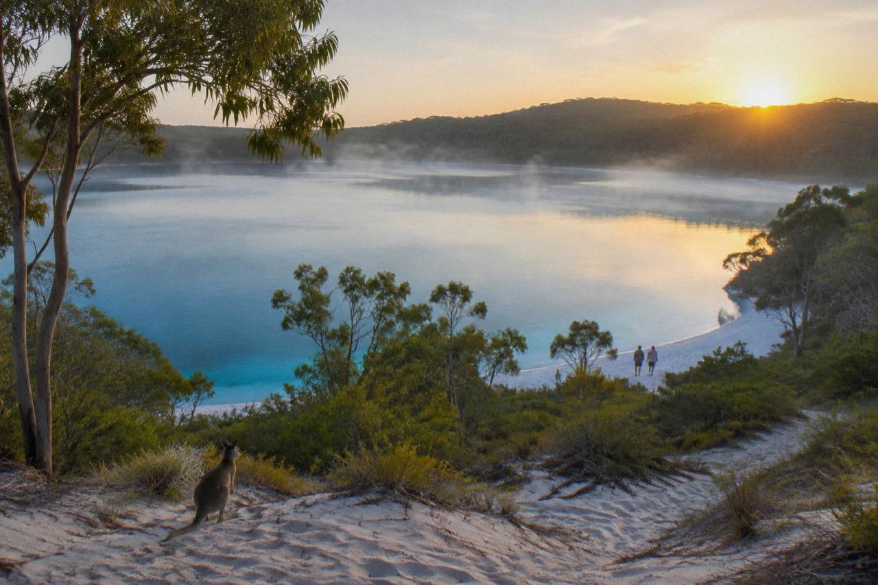 Wide view of a clear freshwater lake ringed by pale sand and bushland on a sand island
