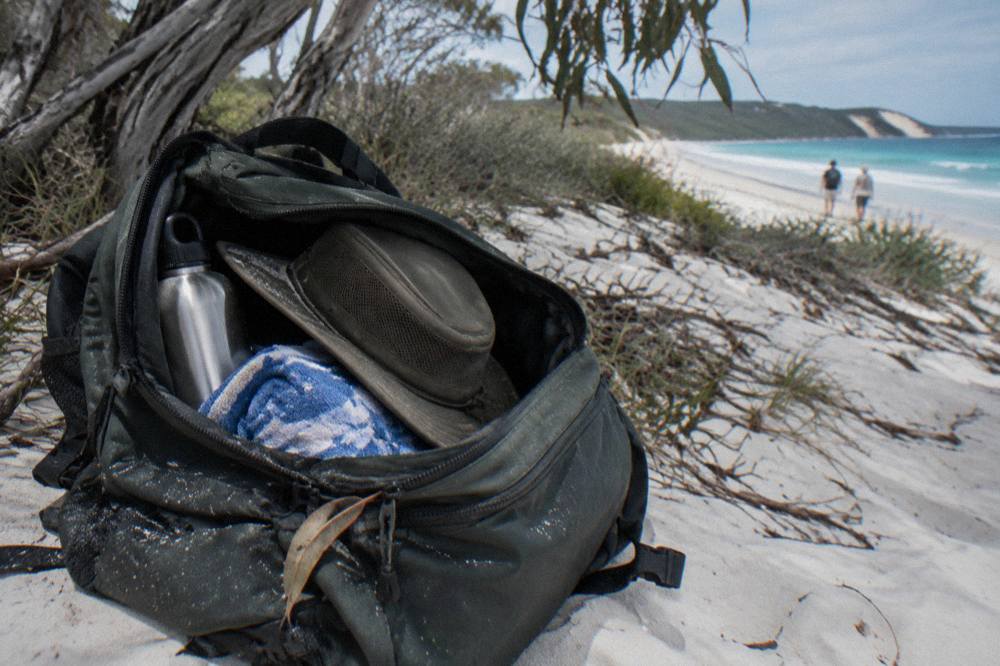 A daypack with a water bottle, hat and towel sitting open on pale sand