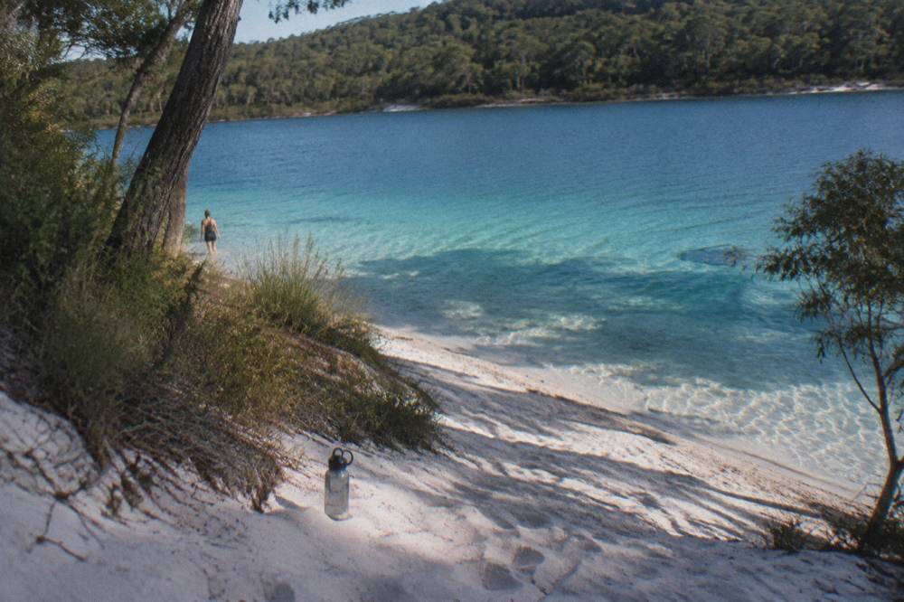 A clear freshwater lake with white sand shore and forest behind it on a calm day