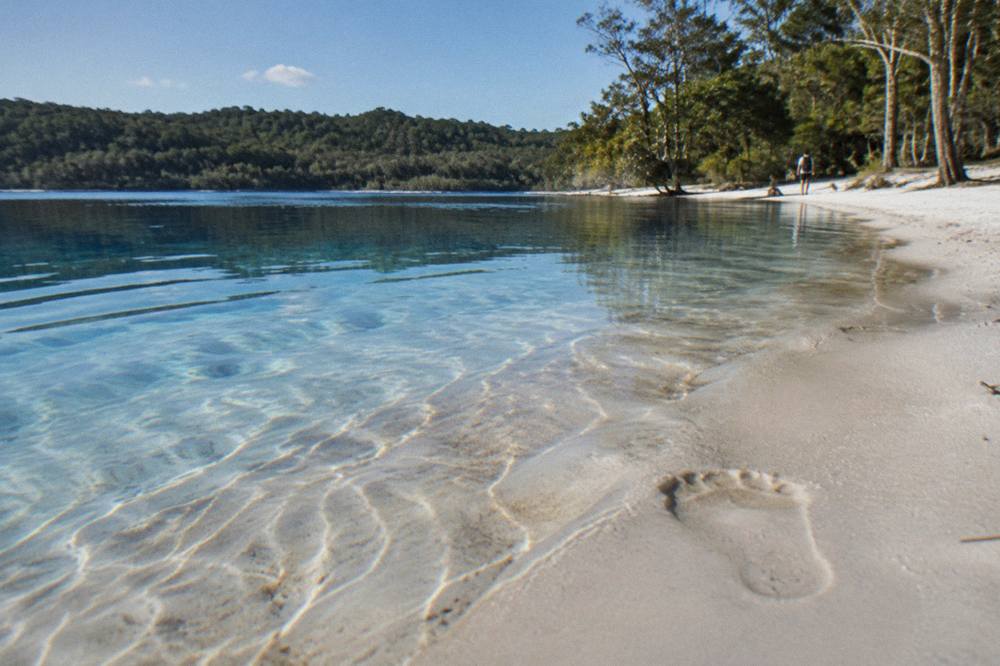 White silica sand shelving into clear blue lake water with forest behind