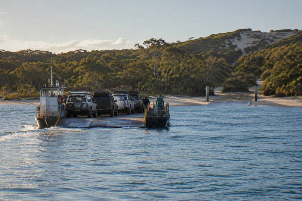 A vehicle barge loaded with four wheel drives crossing calm blue water toward a forested sand island
