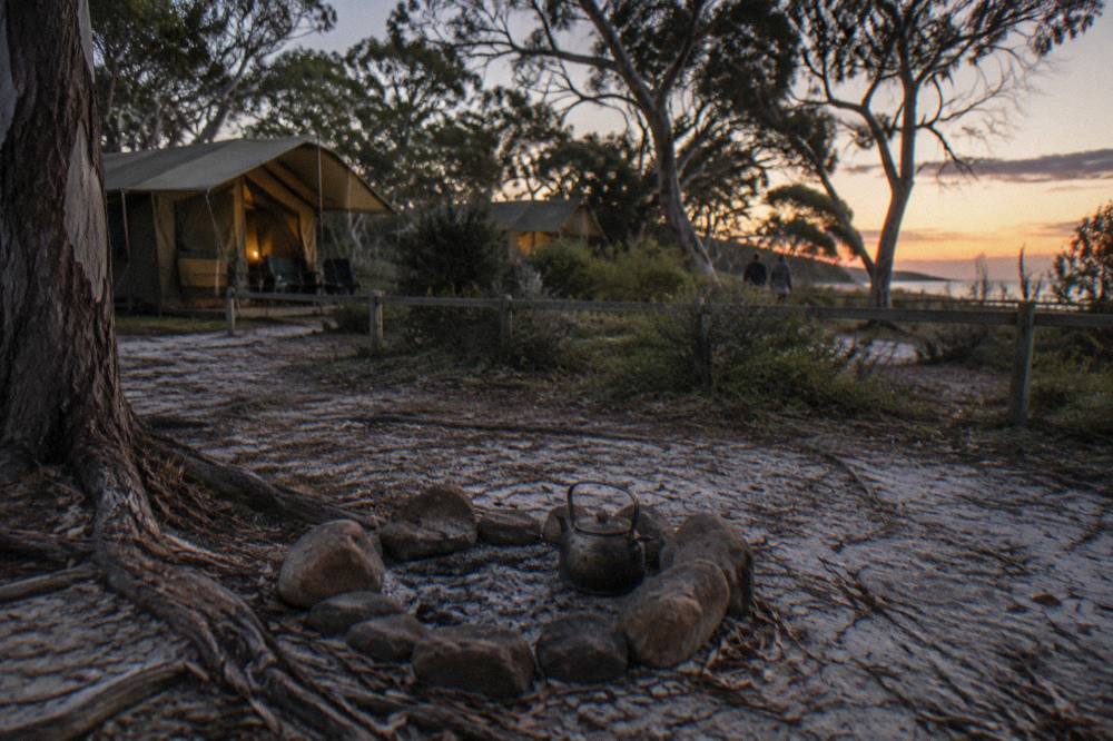 A sandy campsite with permanent canvas tents and a small campfire ring among trees