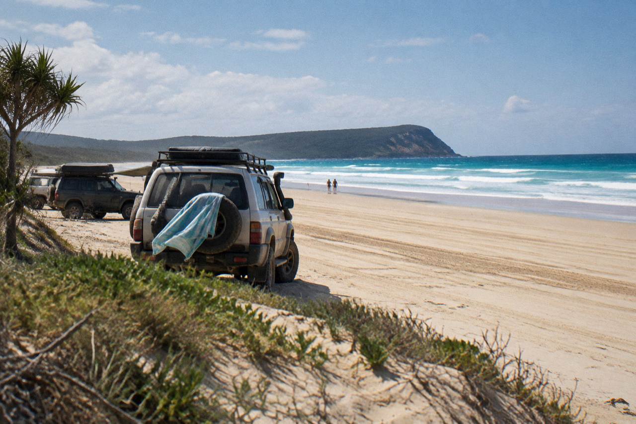 Four wheel drive vehicles parked on a wide sandy beach at K'gari with turquoise water and distant headland