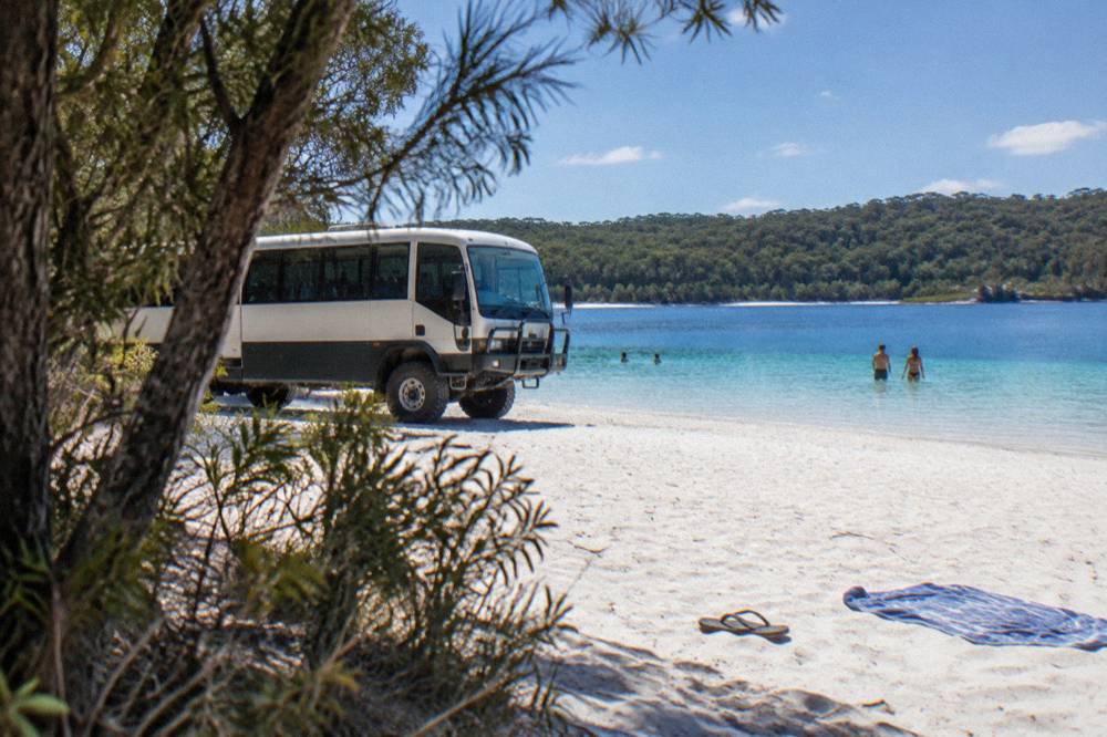 A large four wheel drive coach parked beside a clear freshwater lake ringed by white sand