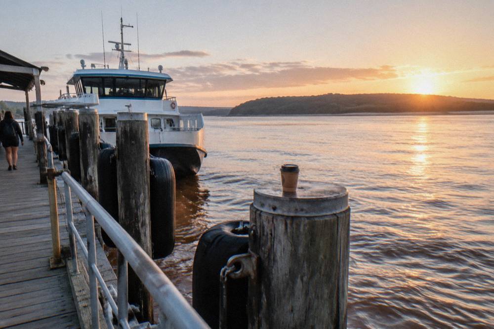A calm bay at sunrise with a ferry vessel at a jetty and low coastal hills across the water