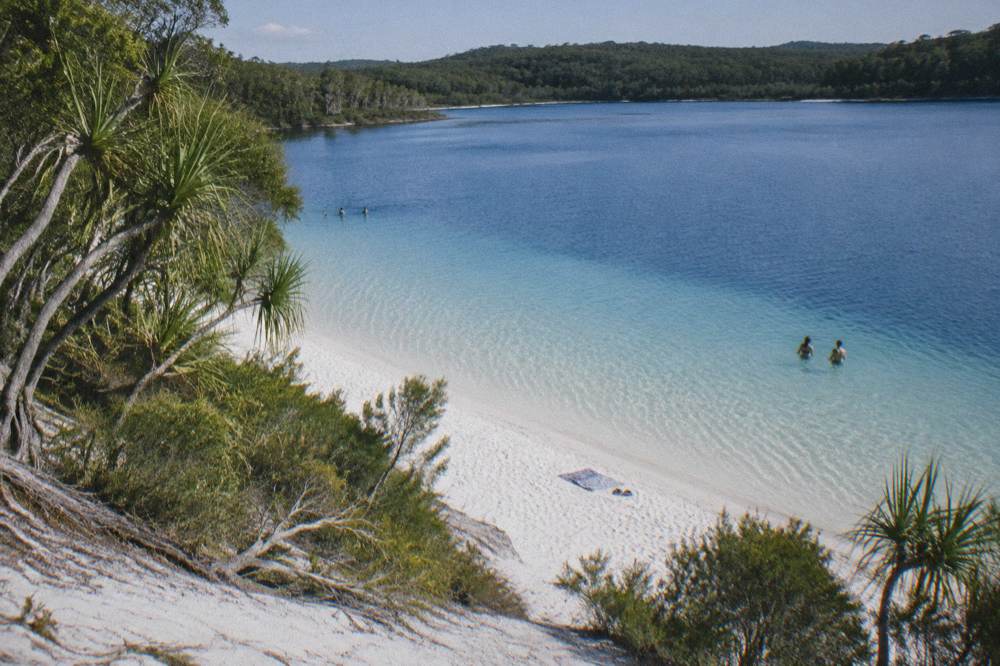 A clear blue freshwater lake ringed by white sand and forest on a sunny day