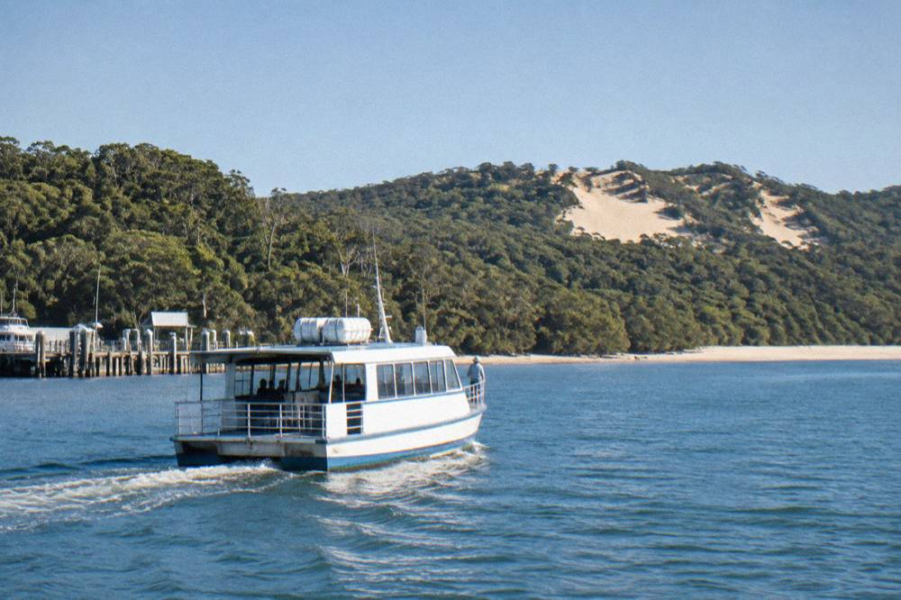 A passenger ferry travelling across calm blue water towards an island shoreline with dense forest