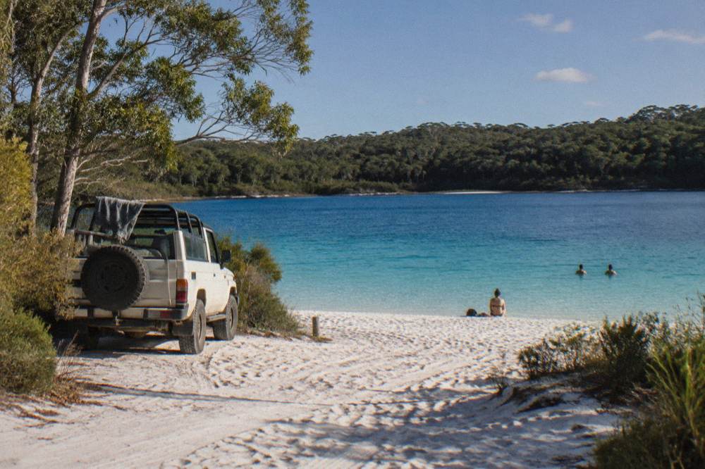 A tour vehicle parked on a wide hard sand beach beside turquoise lake water fringed by trees