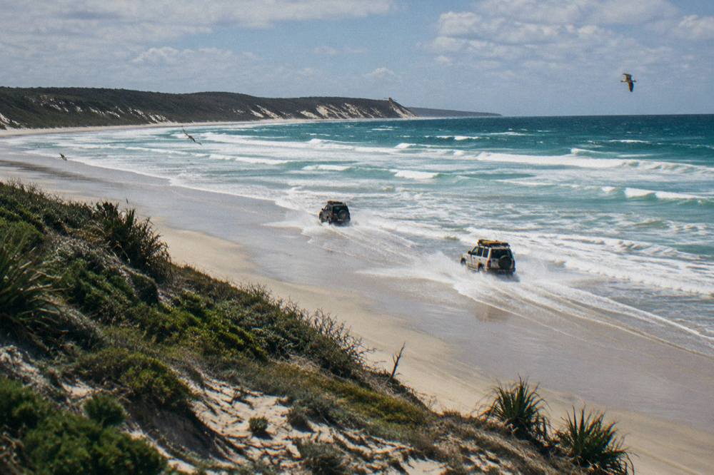 Four wheel drive vehicles travelling along a wide sandy beach beside rolling surf