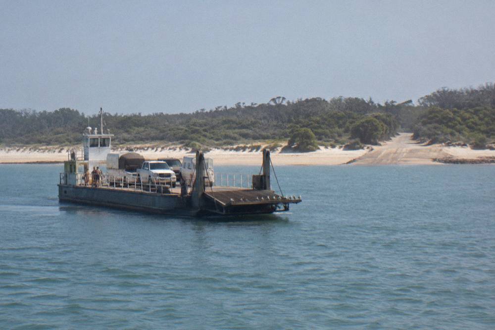 A vehicle barge loaded with four wheel drives crossing calm channel water toward a sand spit