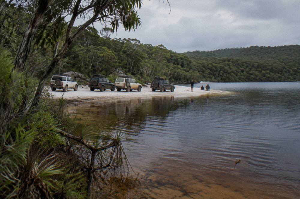 A line of four wheel drives parked on white sand beside a freshwater lake ringed by trees