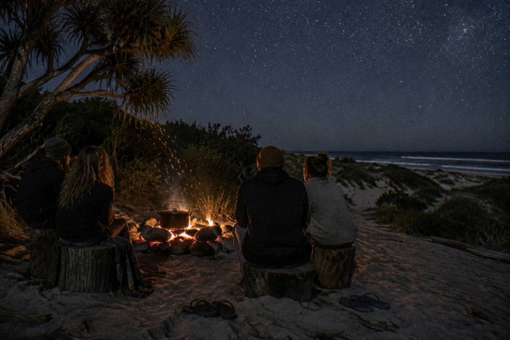 A group of travellers sitting around a campfire on sand under a dark sky