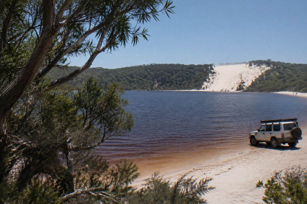 A four wheel drive tour vehicle parked on firm sand beside a freshwater lake surrounded by trees