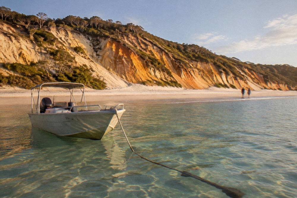 A boat anchored near a remote sandy beach with clear shallow water and tall coloured sand cliffs