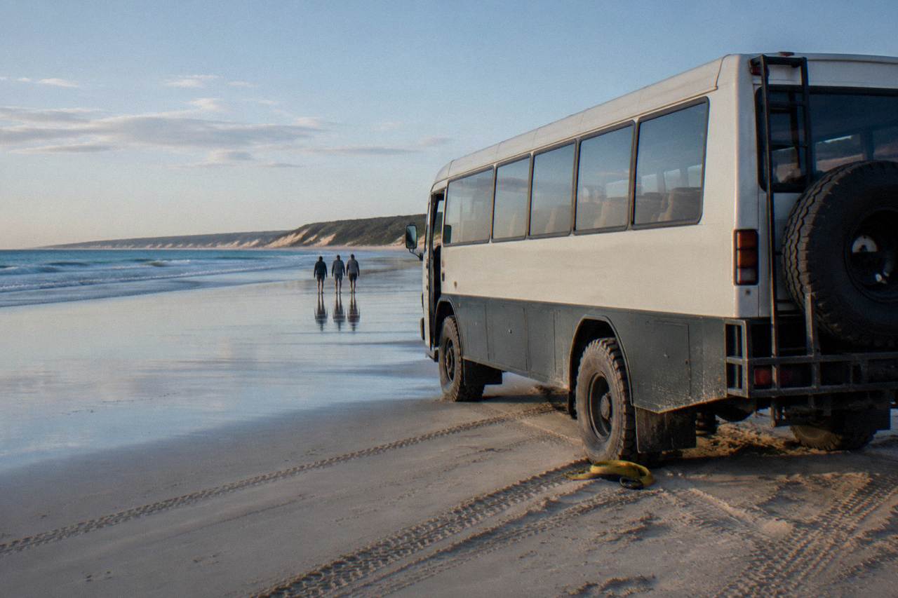 A four wheel drive coach parked on a wide sand beach beside calm blue water