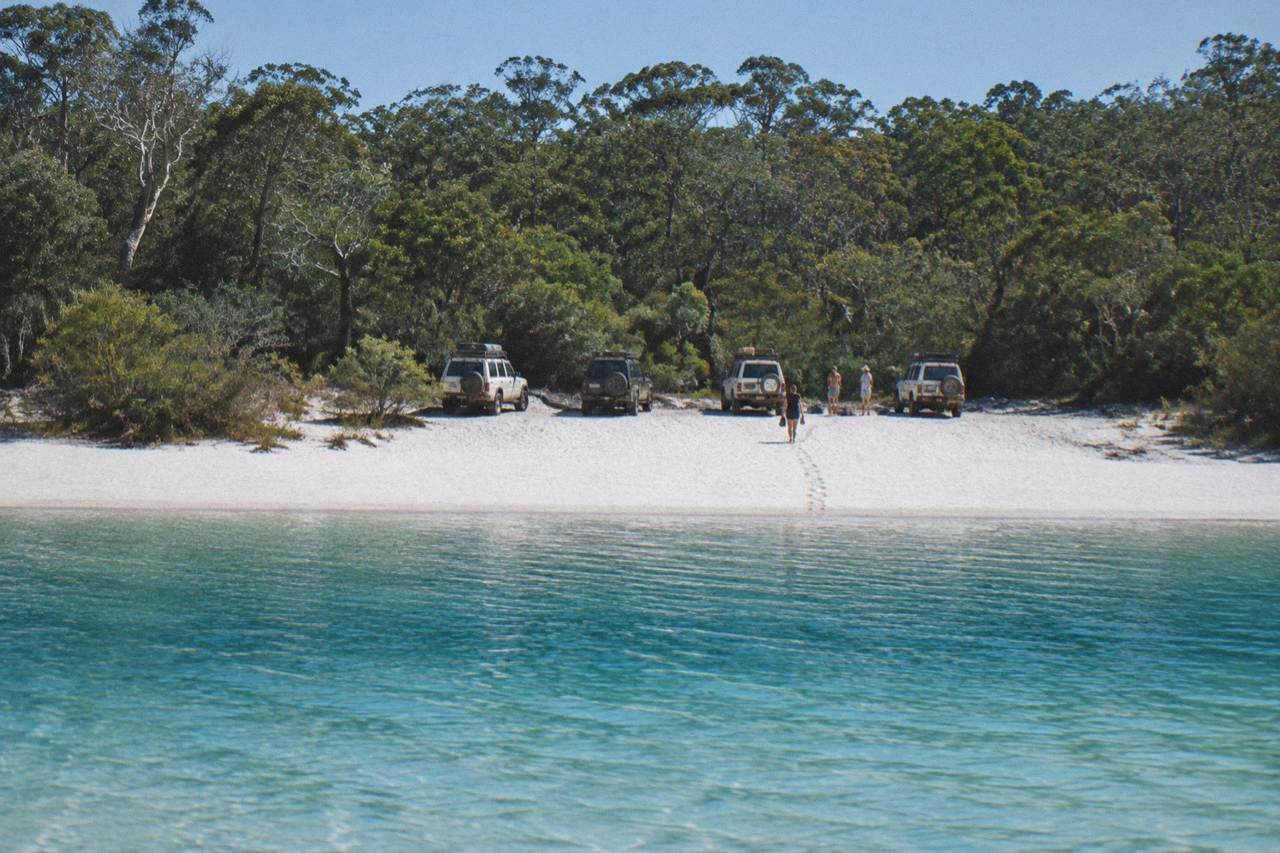 Four wheel drive vehicles parked on a wide sand beach beside turquoise lake water