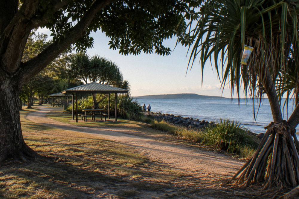 Waterfront parkland path with shade trees and benches beside a calm bay
